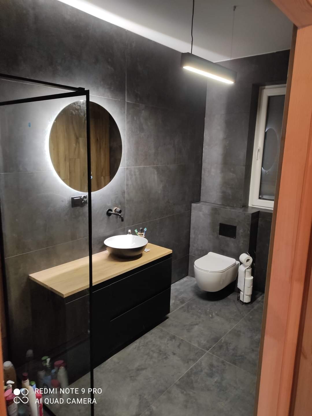 Nowoczesna łazienka z szarymi płytkami, okrągłym lustrem z podświetleniem LED, czarną szafką z drewnianym blatem i wiszącą toaletą. Minimalistyczny design.