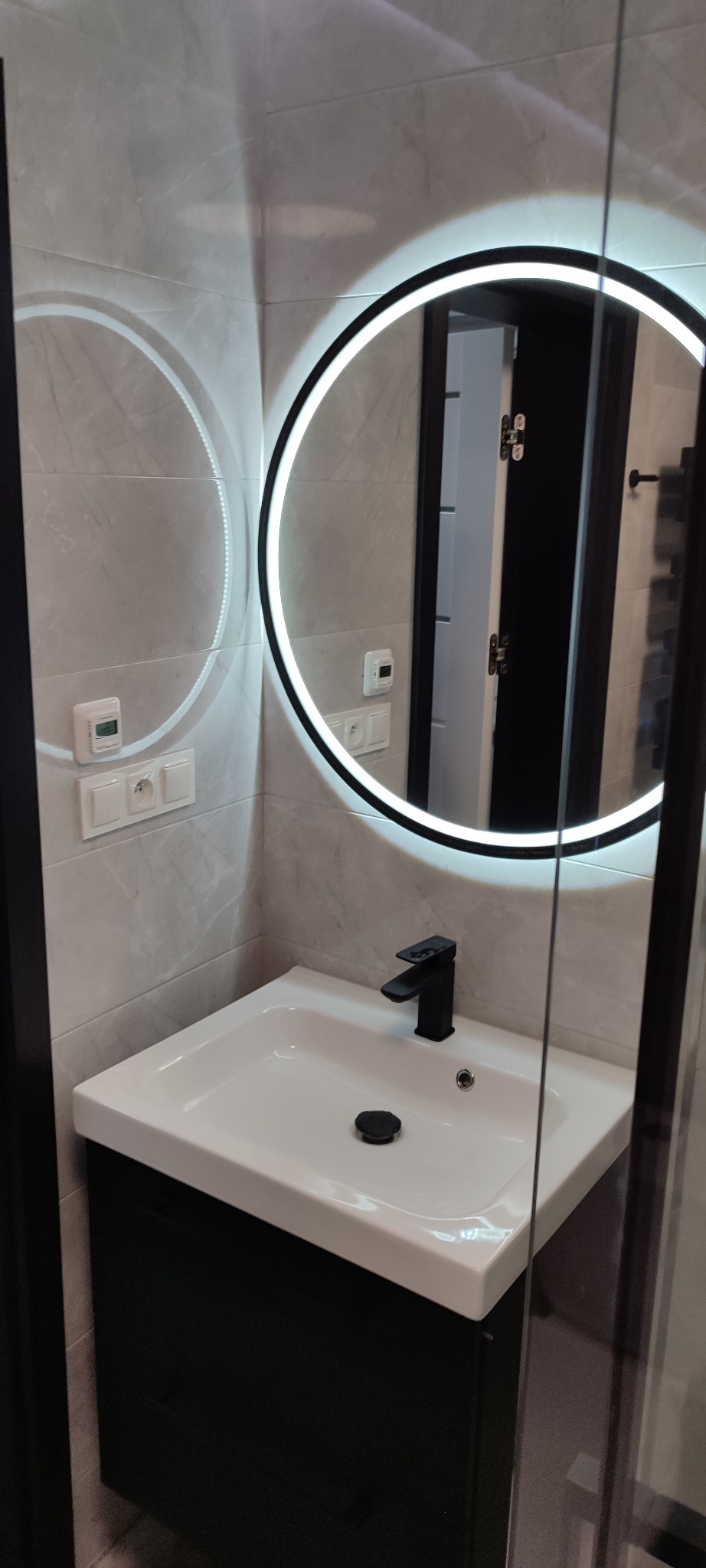 Nowoczesna łazienka: biała umywalka z czarną armaturą, okrągłe lustro z podświetleniem LED na tle szarych płytek. Minimalistyczny design.