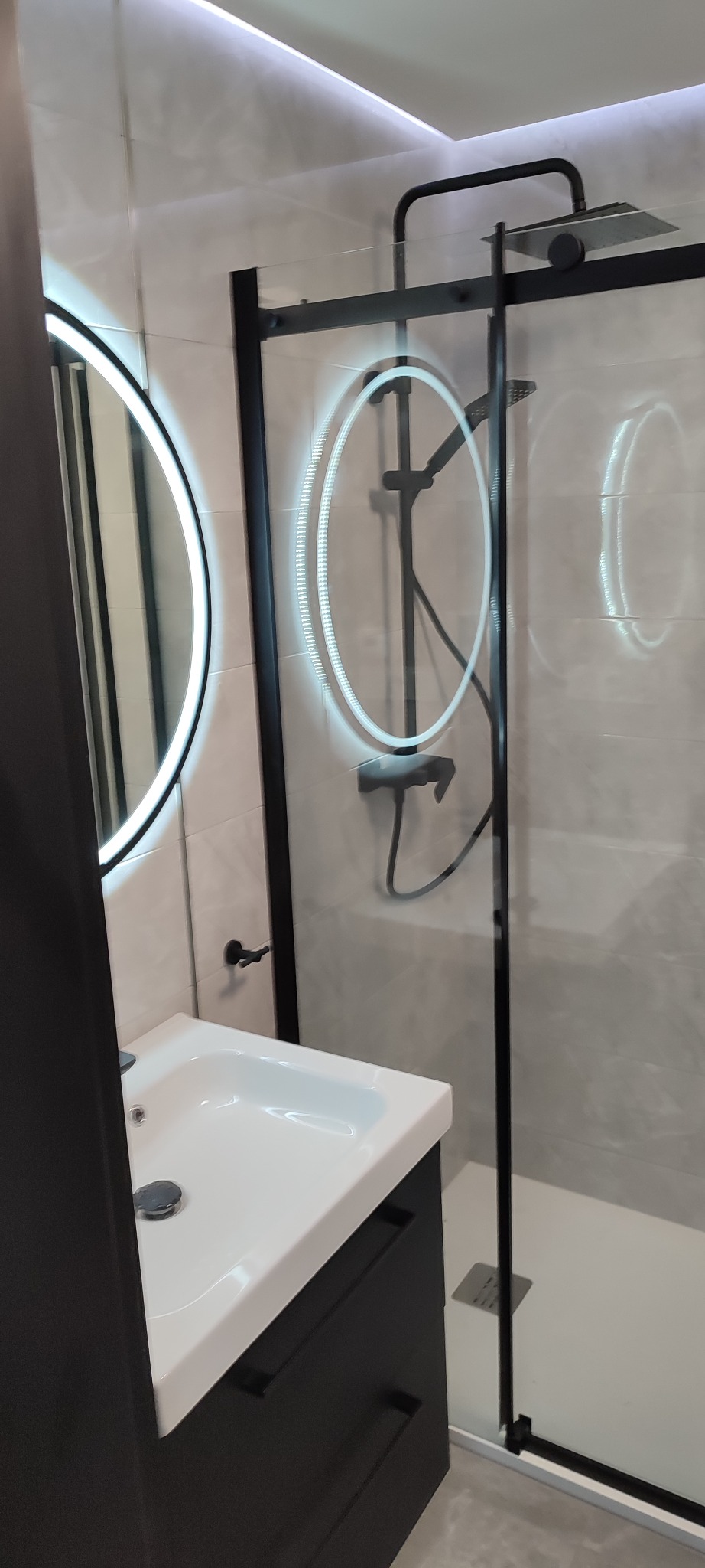 Nowoczesna łazienka z czarnymi akcentami: okrągłe lustro z podświetleniem LED, kabina prysznicowa z czarną ramą i biała umywalka z szafką.