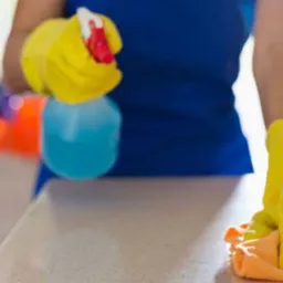 Osoba w ż&oacute;łtych rękawicach czyści blat płynem w spryskiwaczu i pomarańczową ściereczką. W tle rozmyte wnętrze mieszkania.