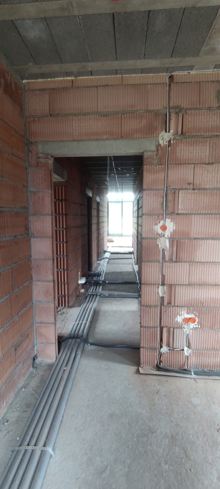 Wnętrze w trakcie prac: korytarz z widoczną instalacją elektryczną w peszlach na podłodze i puszkami w ścianach z czerwonej cegły. Stan surowy.