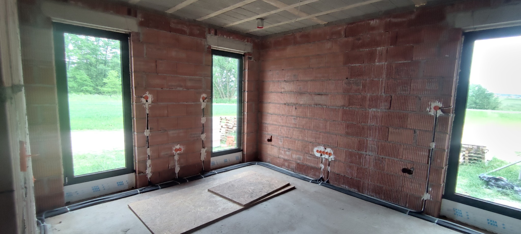 Wnętrze w trakcie prac: mury z cegły, okna z widokiem na zieleń, widoczne kable i puszki podtynkowe. Etap instalacji przewodów elektrycznych w nowym budynku.