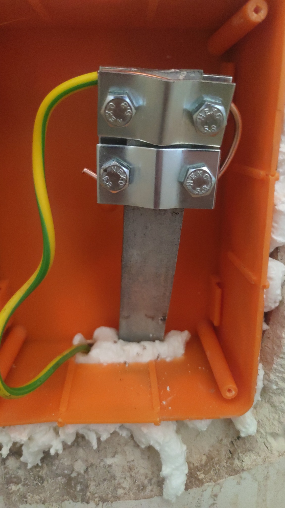 Pomarańczowa puszka elektryczna z metalową szyną i przewodem ochronnym, zabezpieczona pianką montażową. Widok z bliska na połączenie.