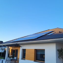 Glow Eco Aleksander Milachowski - Dom jednorodzinny z panelami słonecznymi na dachu w słoneczny dzień. Widoczny ogród i taras z meblami.