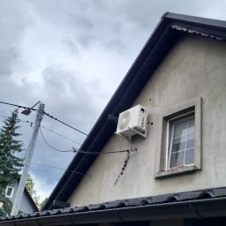 Glow Eco Aleksander Milachowski - Klimatyzator na elewacji domu jednorodzinnego, widok z dołu, pochmurne niebo w tle. Antena satelitarna obok okna. Widoczne kable i fragment drabiny.