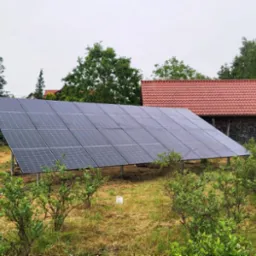 Panel fotowoltaiczny na łące, widok z dołu. W tle budynek z czerwoną dachówką i drzewa. Po lewej drabina. Pochmurne niebo, naturalne oświetlenie.
