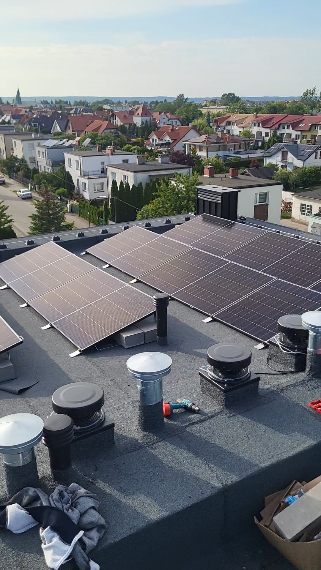 Panele fotowoltaiczne na dachu budynku mieszkalnego, widok z góry, miasto w tle. Na dachu widoczne narzędzia.