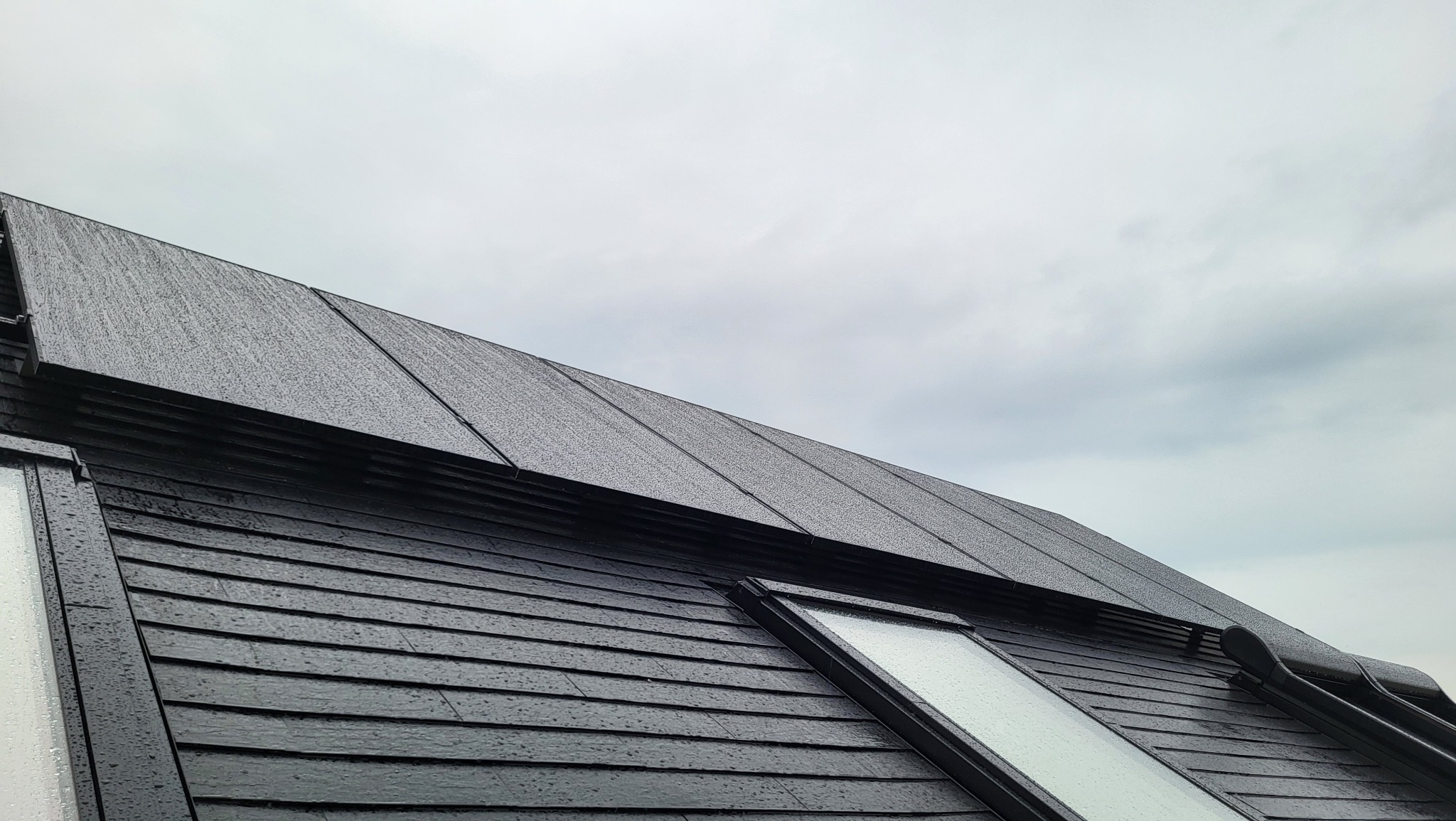 Czarny dach z panelami fotowoltaicznymi i oknem dachowym w deszczowy dzień, widok z dołu na pochmurne niebo.
