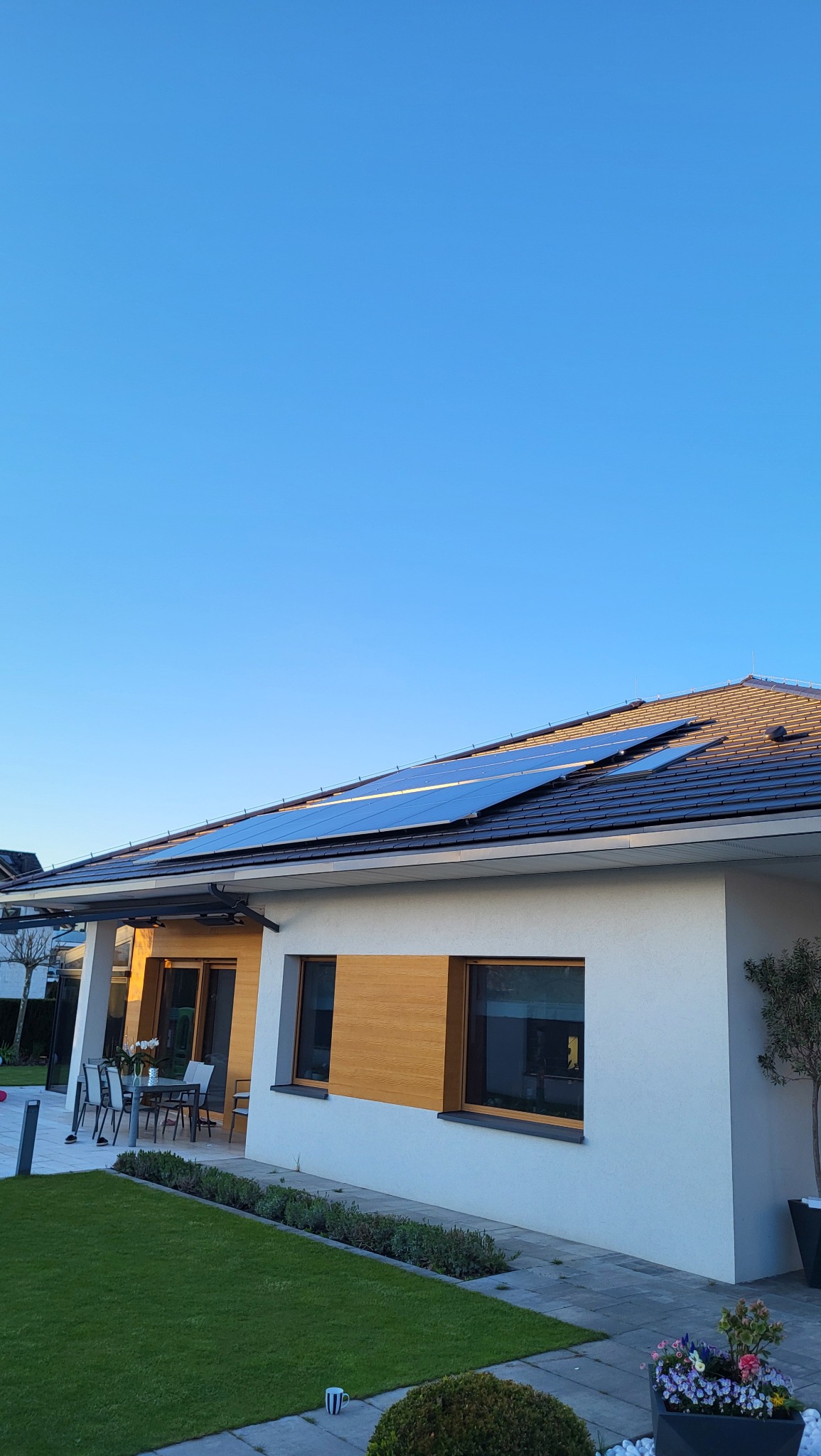 Dom jednorodzinny z panelami słonecznymi na dachu w słoneczny dzień. Widoczny ogród i taras z meblami.