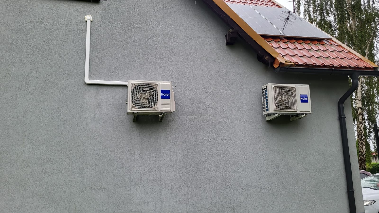 Dwa klimatyzatory Haier na szarej ścianie domu z panelami słonecznymi na dachu. Jeden klimatyzator z rurą odprowadzającą, drugi obok rynny.