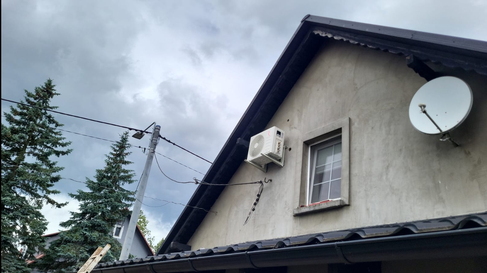 Klimatyzator na elewacji domu jednorodzinnego, widok z dołu, pochmurne niebo w tle. Antena satelitarna obok okna. Widoczne kable i fragment drabiny.