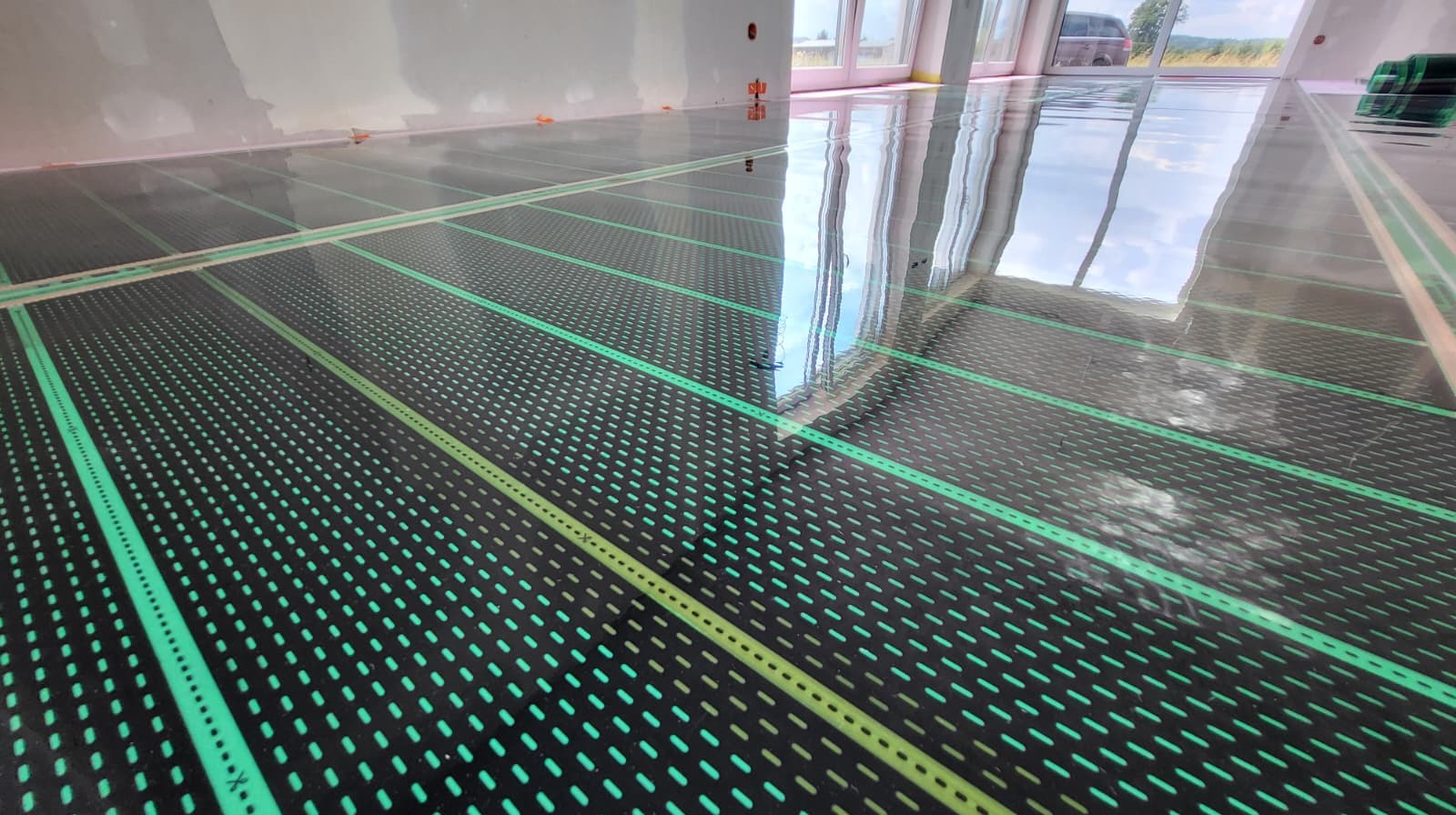 Rozłożone maty grzewcze na podłodze w jasnym pomieszczeniu z oknami. Widoczne odbicie okna w połyskującej powierzchni mat.