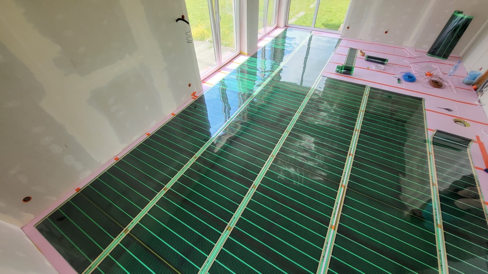 Instalacja mat grzewczych na podłodze w jasnym pomieszczeniu z dużymi oknami, widoczne narzędzia i materiały montażowe. Etap prac przed wylaniem posadzki.