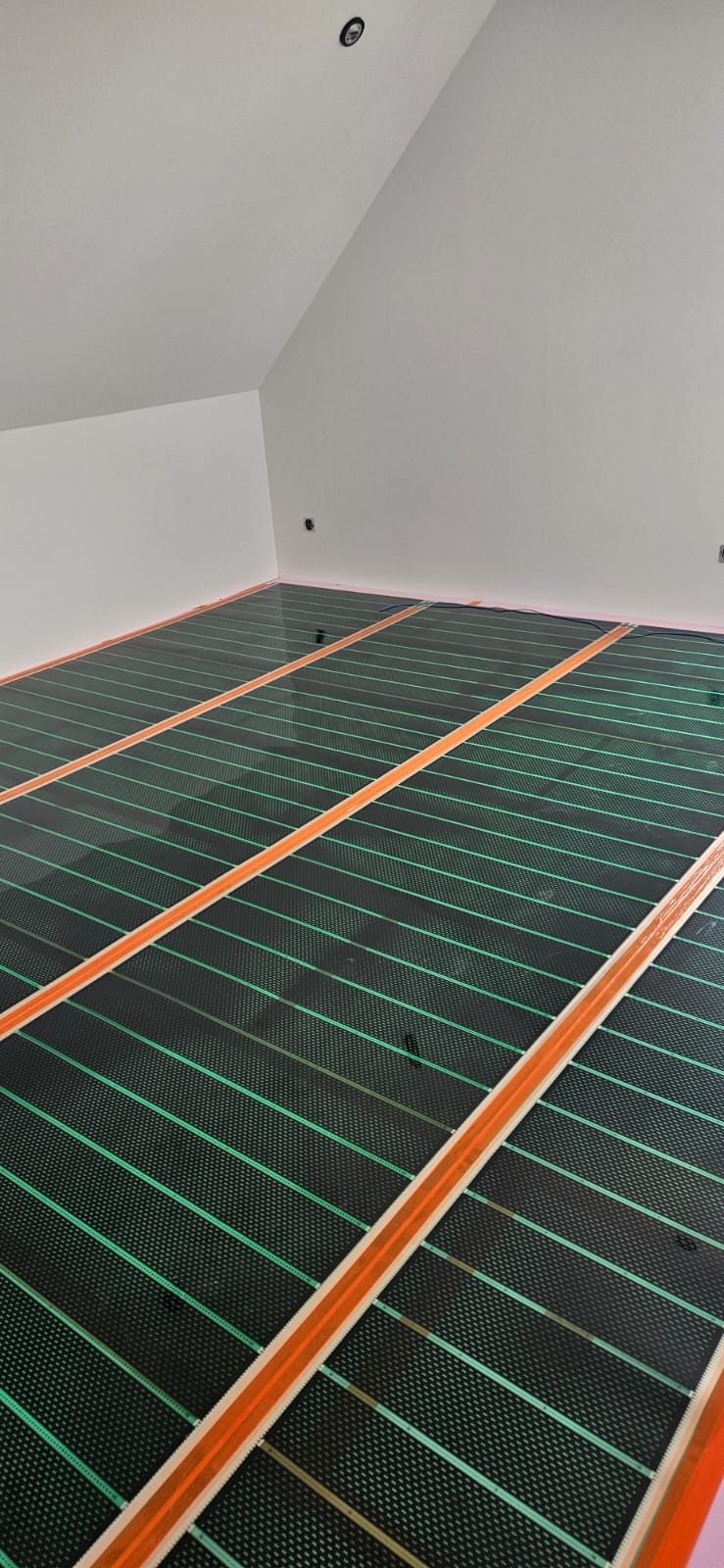 Montaż maty grzewczej na poddaszu. Widoczne równoległe pasy grzewcze w kolorze zielonym, oddzielone pomarańczowymi taśmami. Białe ściany i skośny sufit.