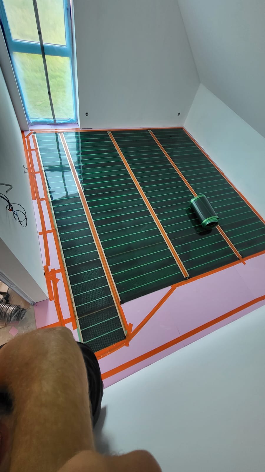Instalacja maty grzewczej na poddaszu: zielona mata z pomarańczowymi paskami, różowa izolacja, okno zabezpieczone folią. Widok z góry, fragment ciała instalatora w kadrze.