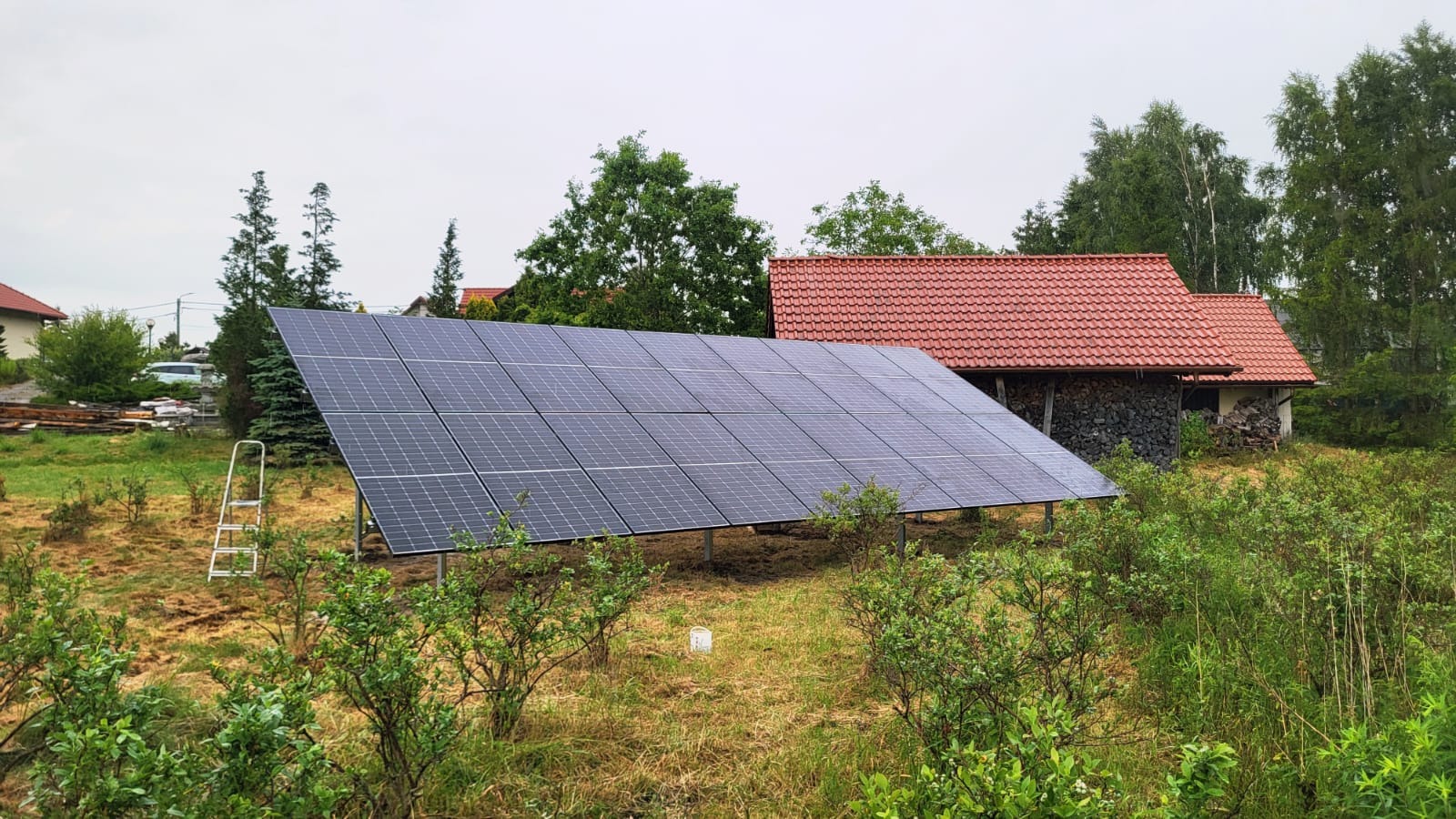 Panel fotowoltaiczny na łące, widok z dołu. W tle budynek z czerwoną dachówką i drzewa. Po lewej drabina. Pochmurne niebo, naturalne oświetlenie.