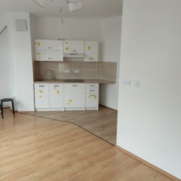 Adam Nowak - Jasna kuchnia z drewnianą podłogą w Konstancin-Jeziorna. Białe szafki z naklejkami zabezpieczającymi. Widoczny fragment podłogi i krzesło.