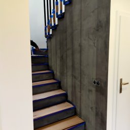 Adam Nowak - Klatka schodowa w trakcie malowania: szare ściany z efektem dekoracyjnym, zabezpieczone schody i balustrada taśmą malarską. Widoczna dłoń malująca ścianę.