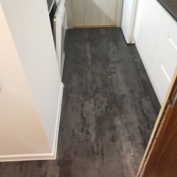 Adam Nowak - Ciemna, teksturowana podłoga w wąskiej kuchni z białymi szafkami i listwami przypodłogowymi. Widoczny fragment drewnianych drzwi i sprzęt AGD.