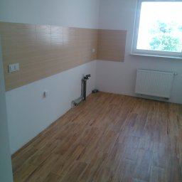 Adam Nowak - Wykończona kuchnia z drewnopodobną podłogą i częściowo położonymi beżowymi płytkami na ścianie. Widoczne przyłącza wodne i grzejnik pod oknem.