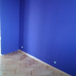 Adam Nowak - Pokój po malowaniu na intensywny, królewski odcień fioletu. Parkiet ułożony w jodełkę, widoczne gniazdka elektryczne. Świeżo odnowione wnętrze.