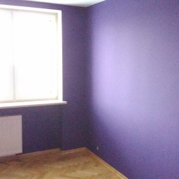 Adam Nowak - Ściana pomalowana na fioletowo z oknem i grzejnikiem. Parkiet ułożony w jodełkę. Widok na róg pokoju z wyraźną strukturą farby na ścianach.