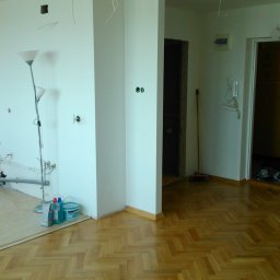 Adam Nowak - Wnętrze pokoju w trakcie remontu: parkiet w jodełkę, białe ściany z instalacjami, lampa i pojemniki z farbą. Częściowo wykończone podłoże pod parkiet.