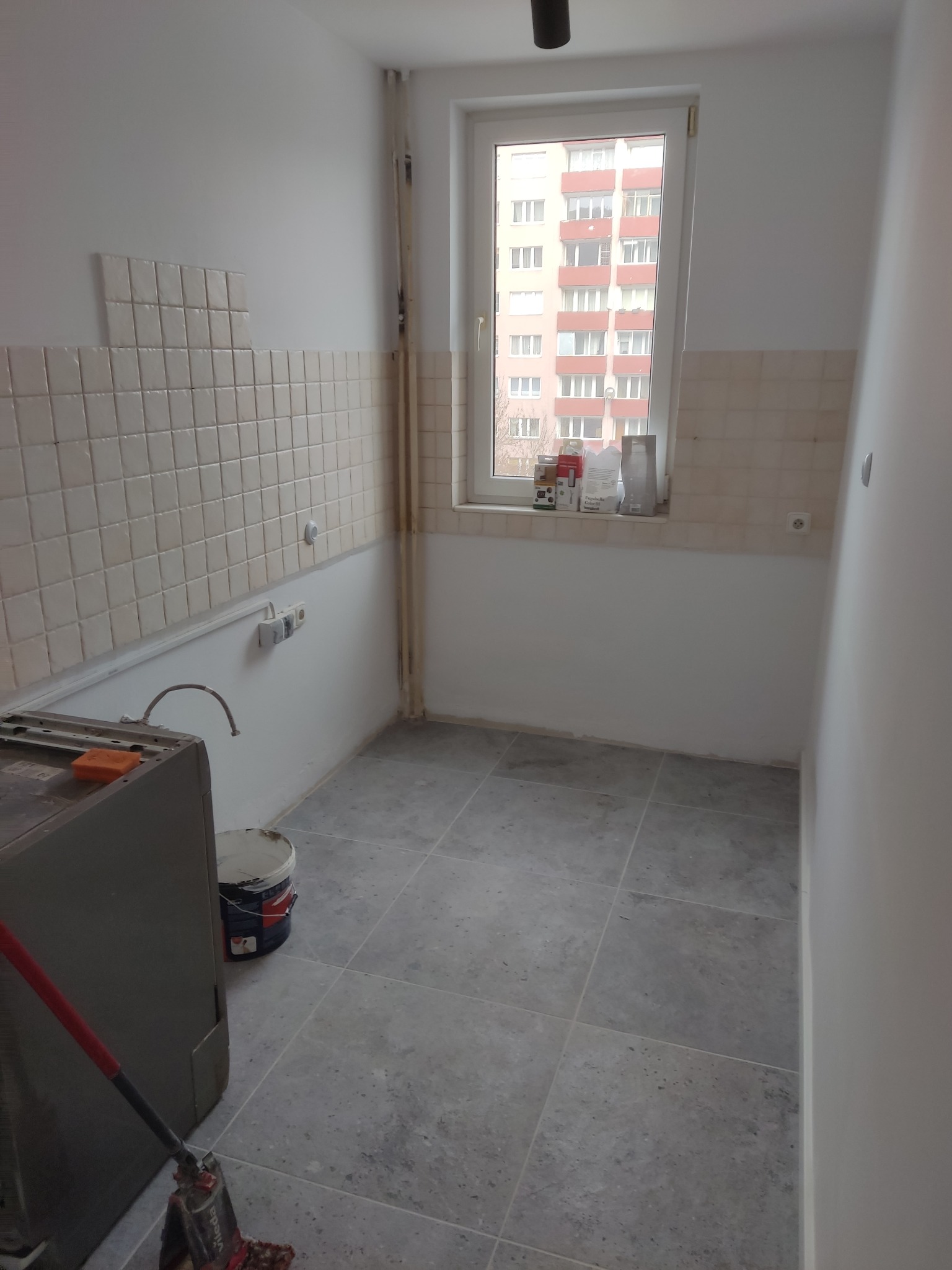 Wnętrze kuchni w trakcie remontu: szare płytki podłogowe, częściowo położone beżowe kafelki na ścianie, wiadro farby i mop w rogu, okno z widokiem na blok.