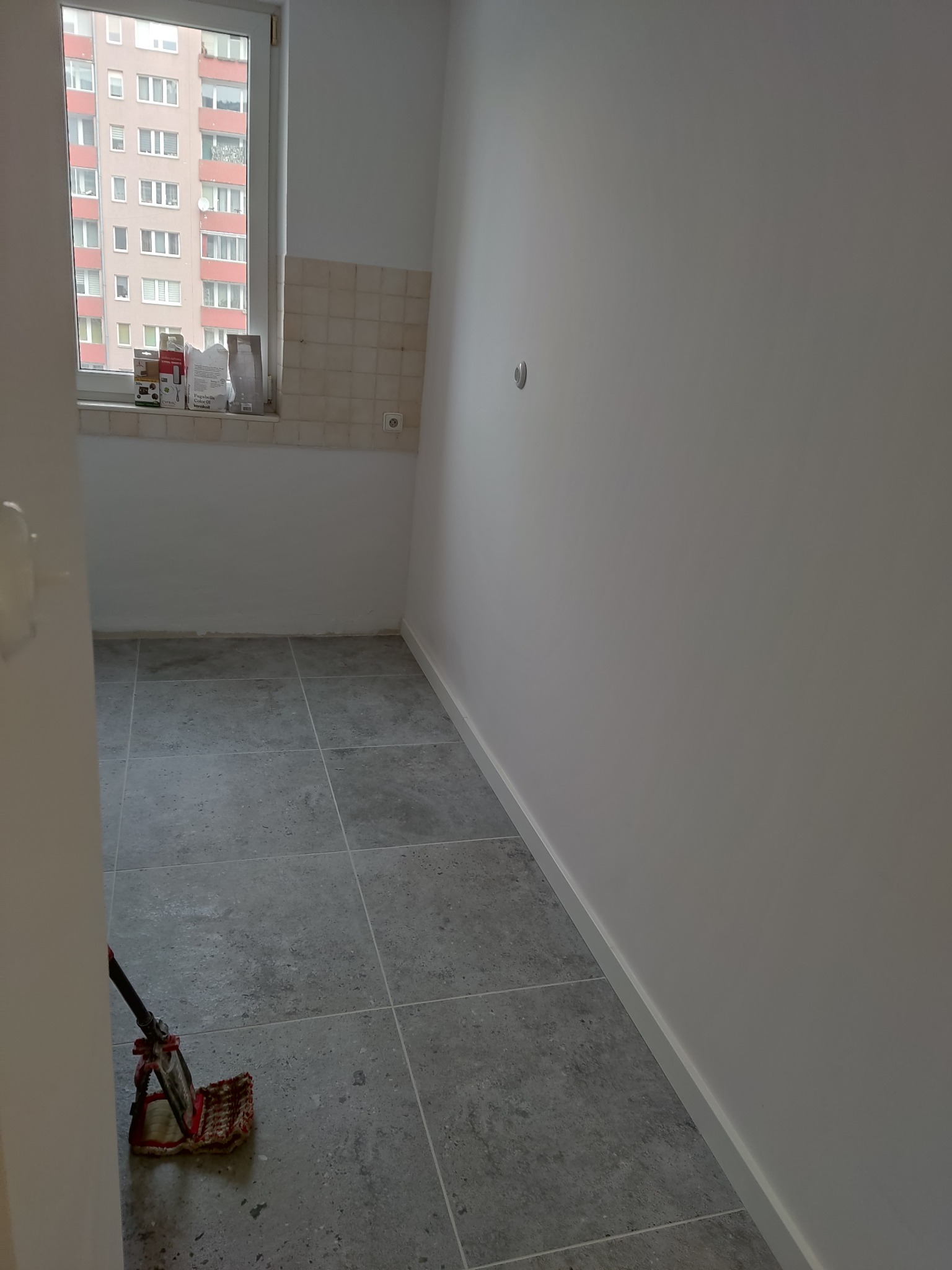 Wnętrze pomieszczenia po remoncie: szare płytki podłogowe, świeżo pomalowane białe ściany z listwami przypodłogowymi, okno z widokiem na budynek.