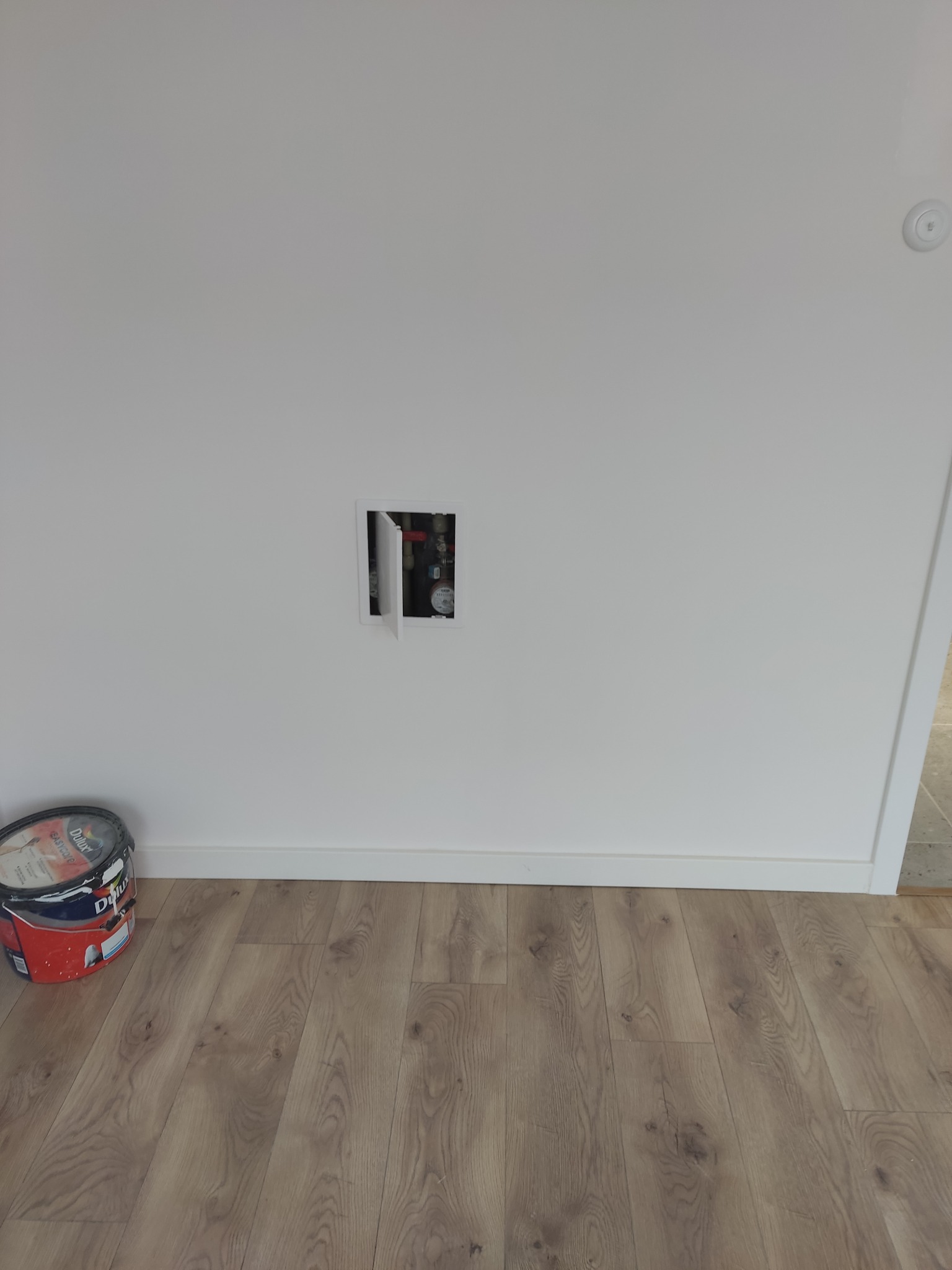 Biała ściana z otworem rewizyjnym, widoczne rury. Na podłodze panele i puszka farby Dulux. Ujęcie z dołu, blisko podłogi. Minimalistyczny kadr.