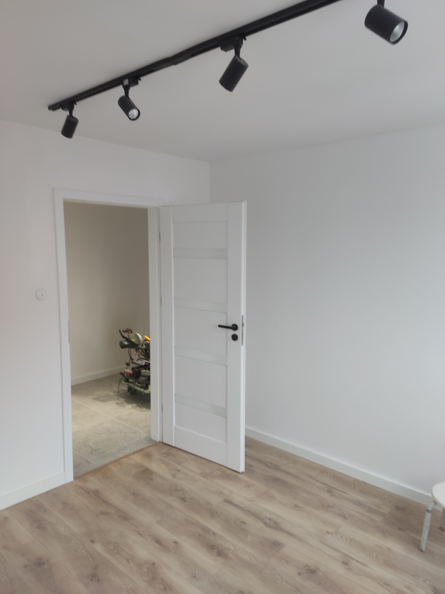 Wnętrze po remoncie: białe ściany, panel podłogowy i białe drzwi z widocznym sprzętem budowlanym w tle. Oświetlenie sufitowe punktowe na szynie.