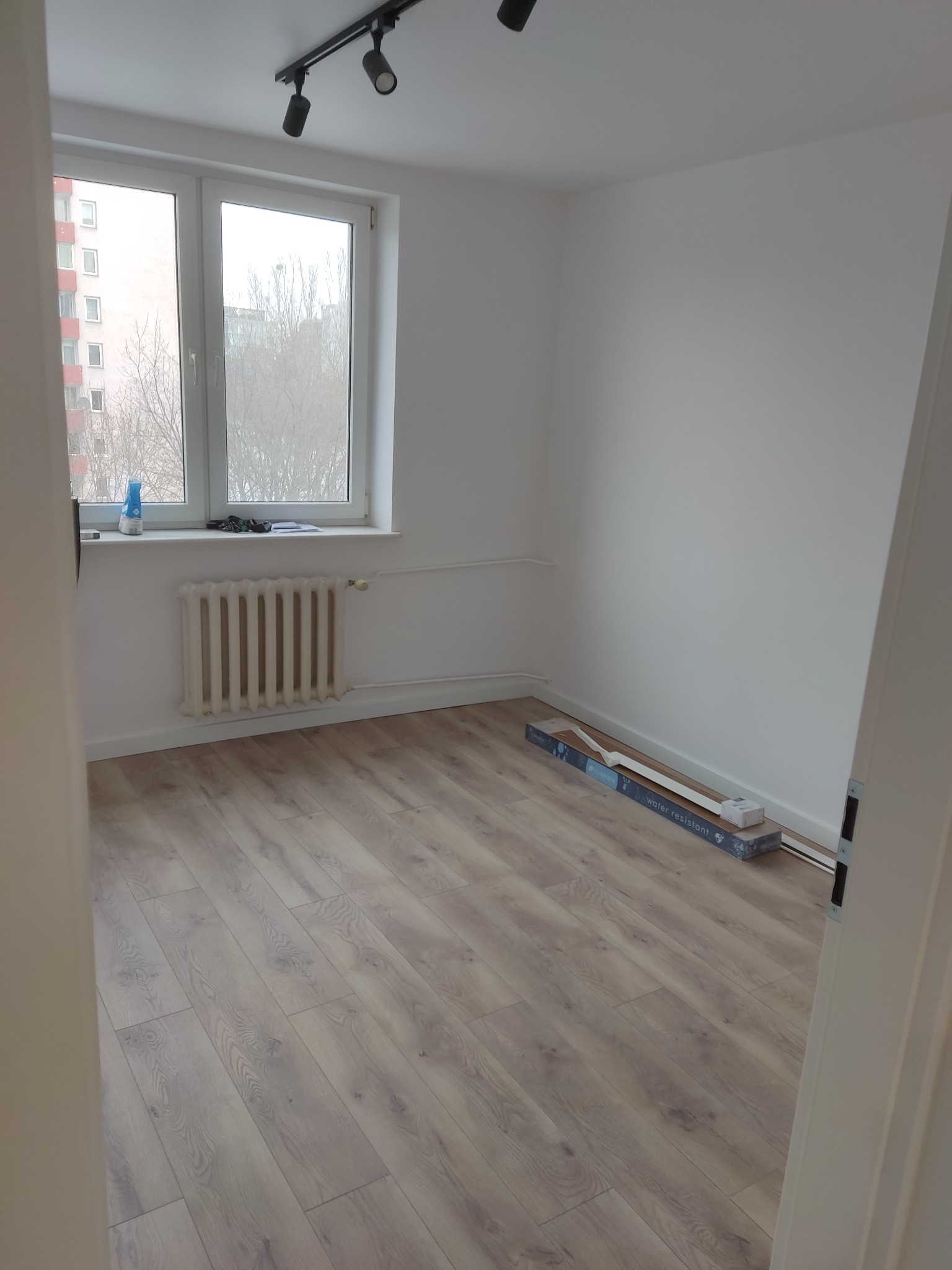 Pokój po remoncie z panelami podłogowymi, grzejnikiem pod oknem i listwami przypodłogowymi w opakowaniu. Widok na budynek zza okna.