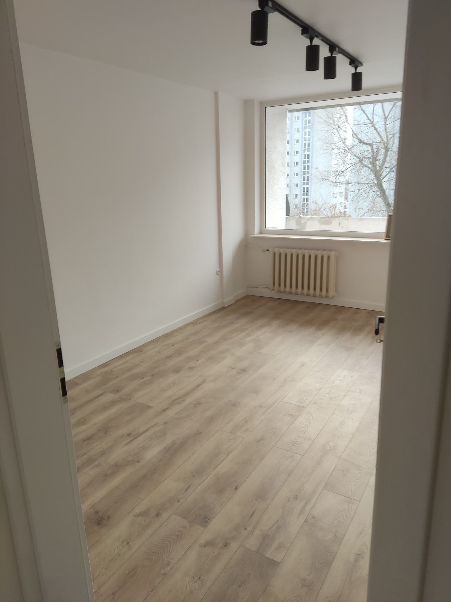 Jasne panele podłogowe w pokoju z białymi ścianami i grzejnikiem pod oknem. Widok zza otwartych drzwi, czarna lampa sufitowa.