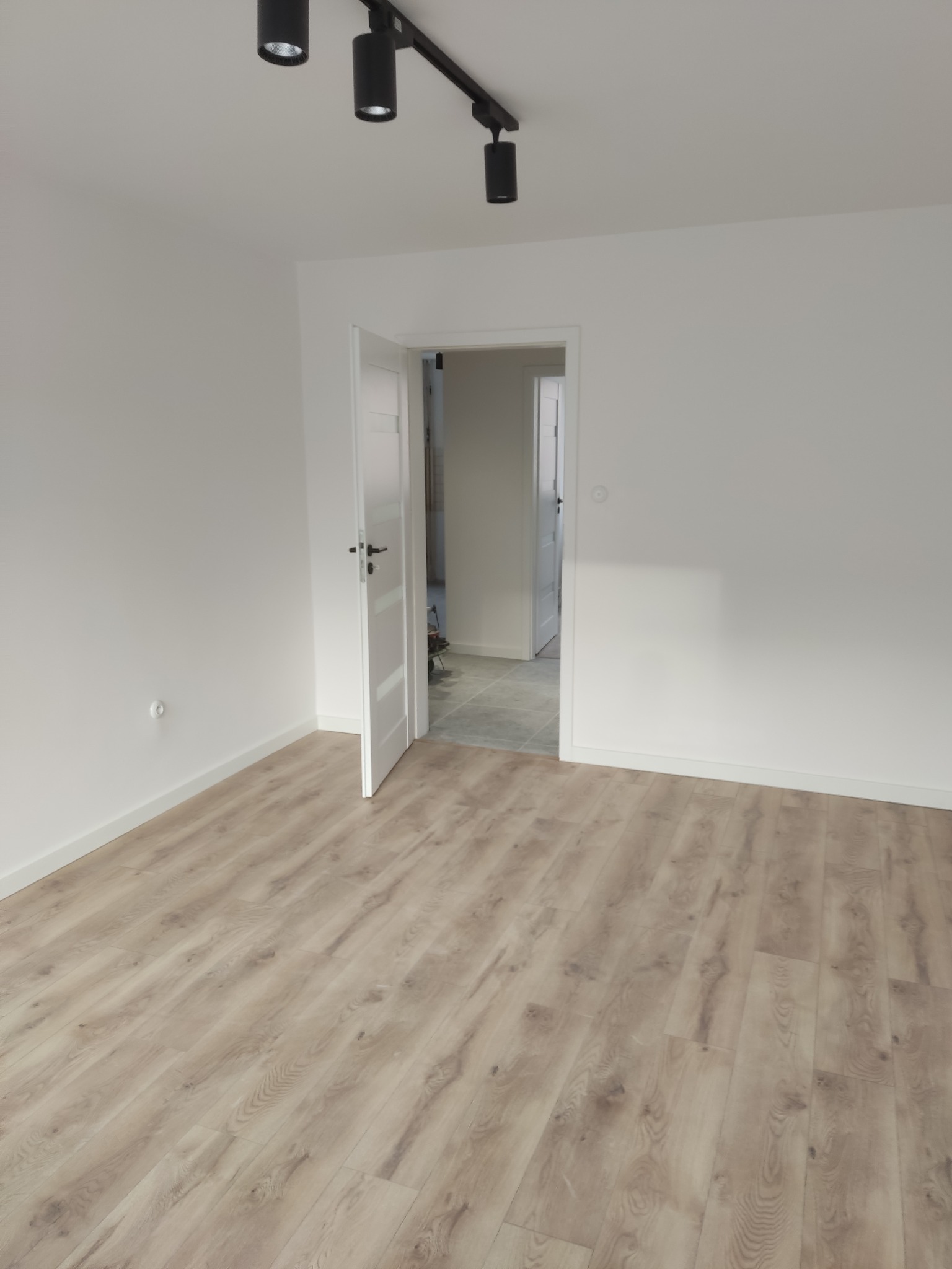 Jasne panele podłogowe w nowym, pomalowanym na biało pokoju z otwartymi, białymi drzwiami. Widoczne czarne reflektory sufitowe.