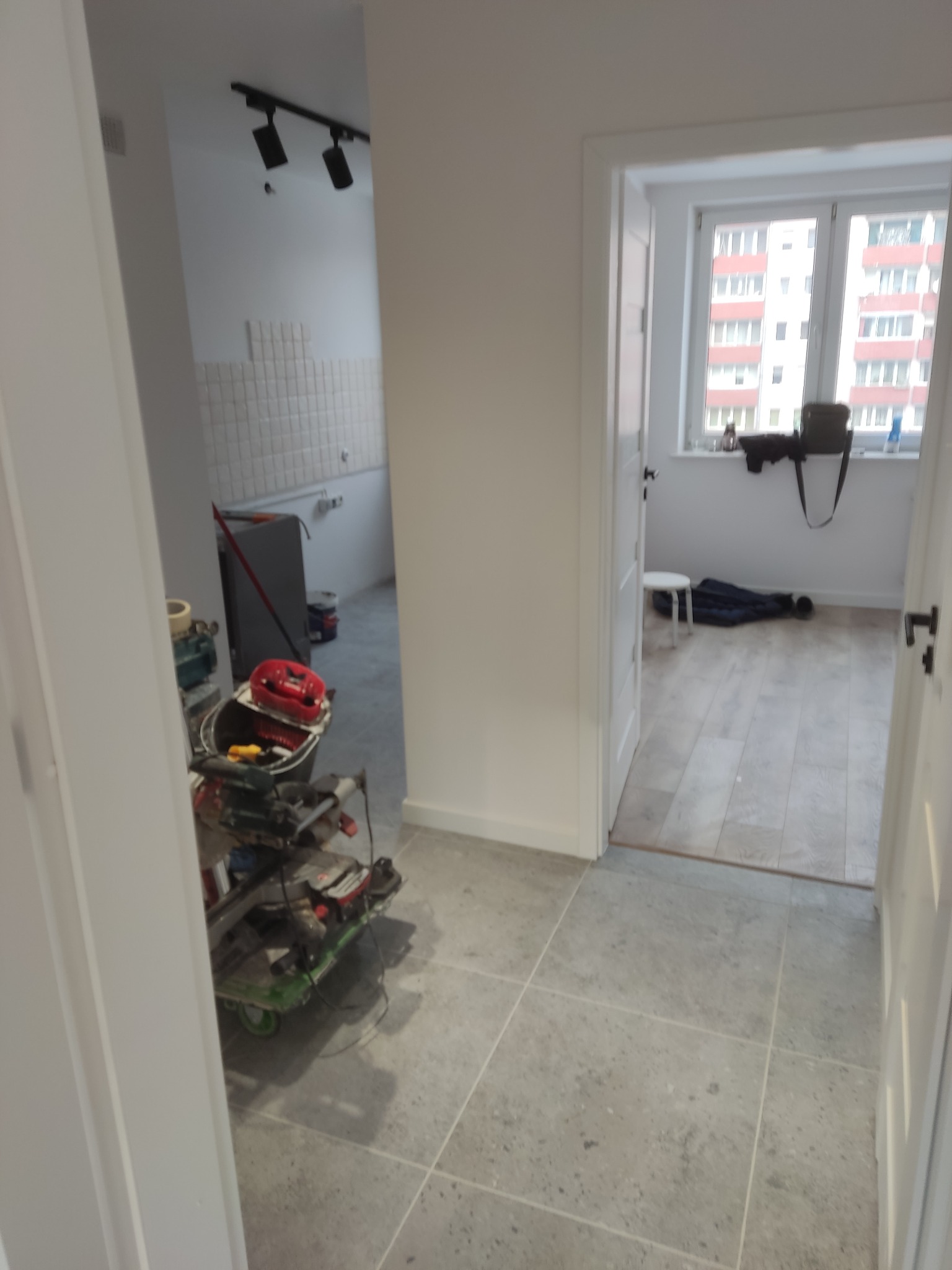Wnętrze w trakcie remontu: szare płytki podłogowe, widoczne narzędzia, fragment ściany z białymi płytkami, okno z widokiem na blok. Remont w toku, bałagan.