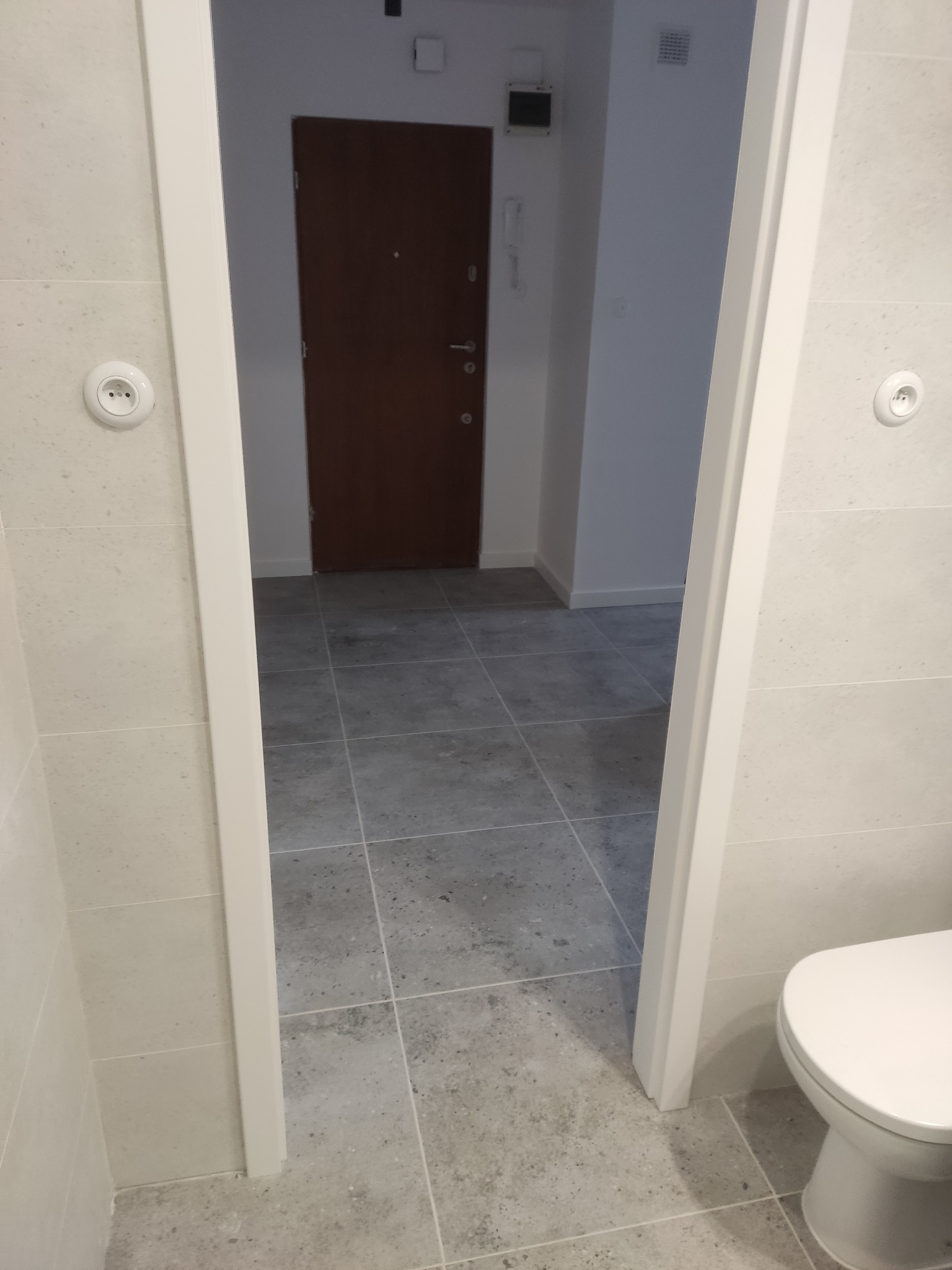 Wykończona łazienka z szarą, kwadratową glazurą podłogową. Widoczna biała toaleta i fragment futryny drzwiowej z gniazdkiem elektrycznym na ścianie.