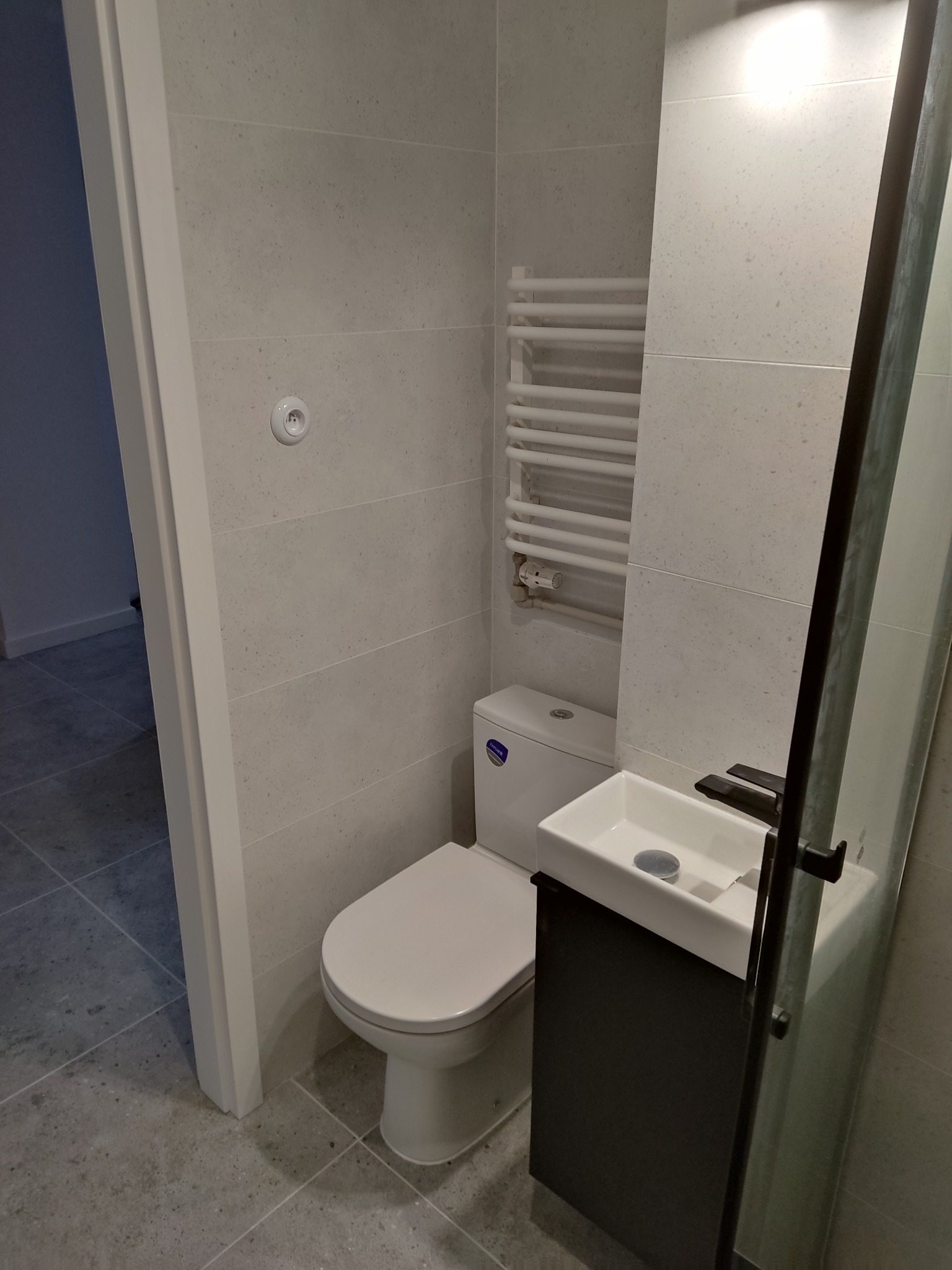 Wykończona łazienka z szarymi płytkami, toaleta, grzejnik drabinkowy i umywalka z szafką. Widok z perspektywy wejścia do pomieszczenia. Minimalistyczny design.