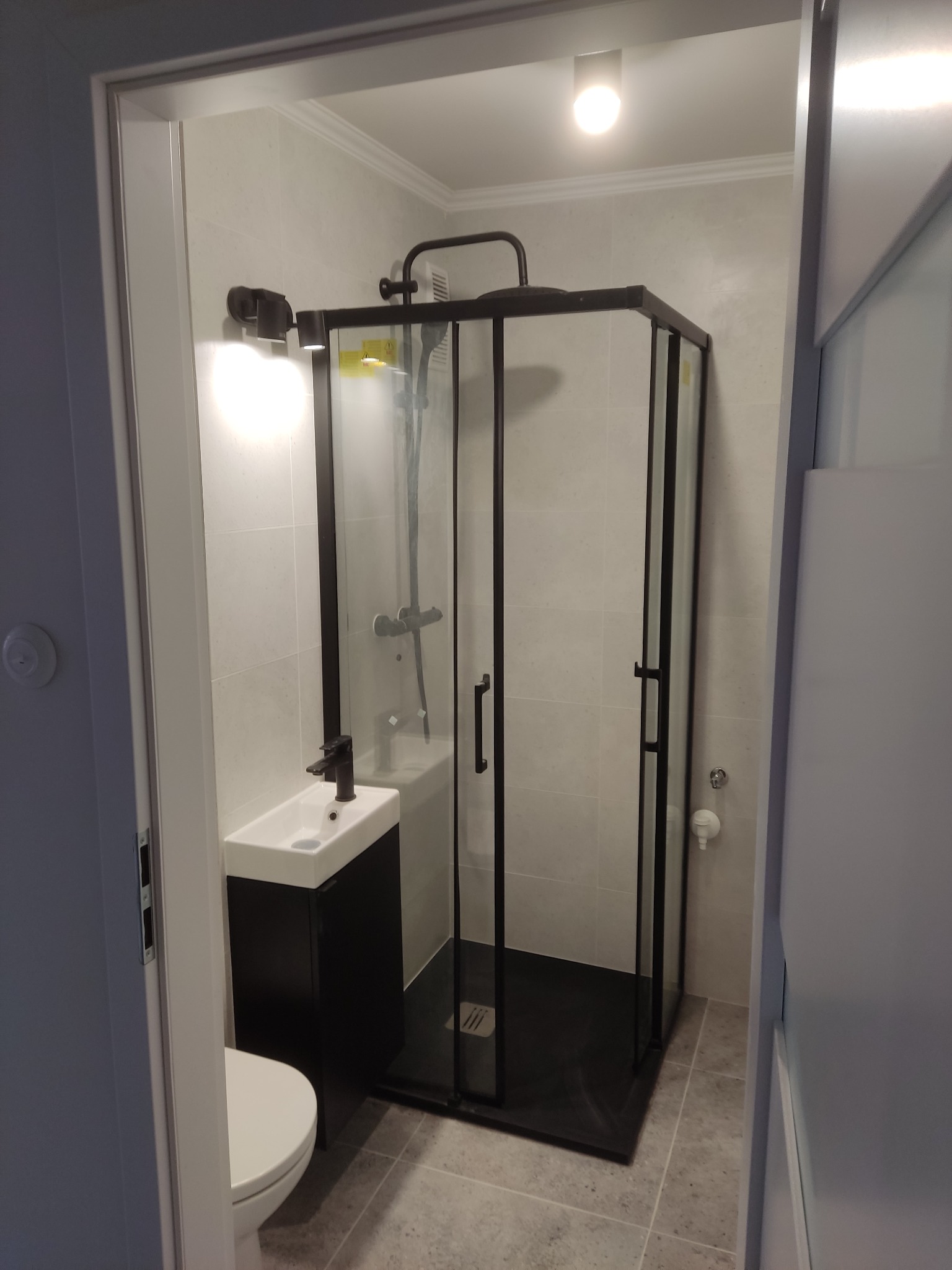 Nowoczesna łazienka z kabiną prysznicową typu walk-in z czarnymi profilami, umywalką na szafce i toaletą. Jasne płytki na ścianach i podłodze.