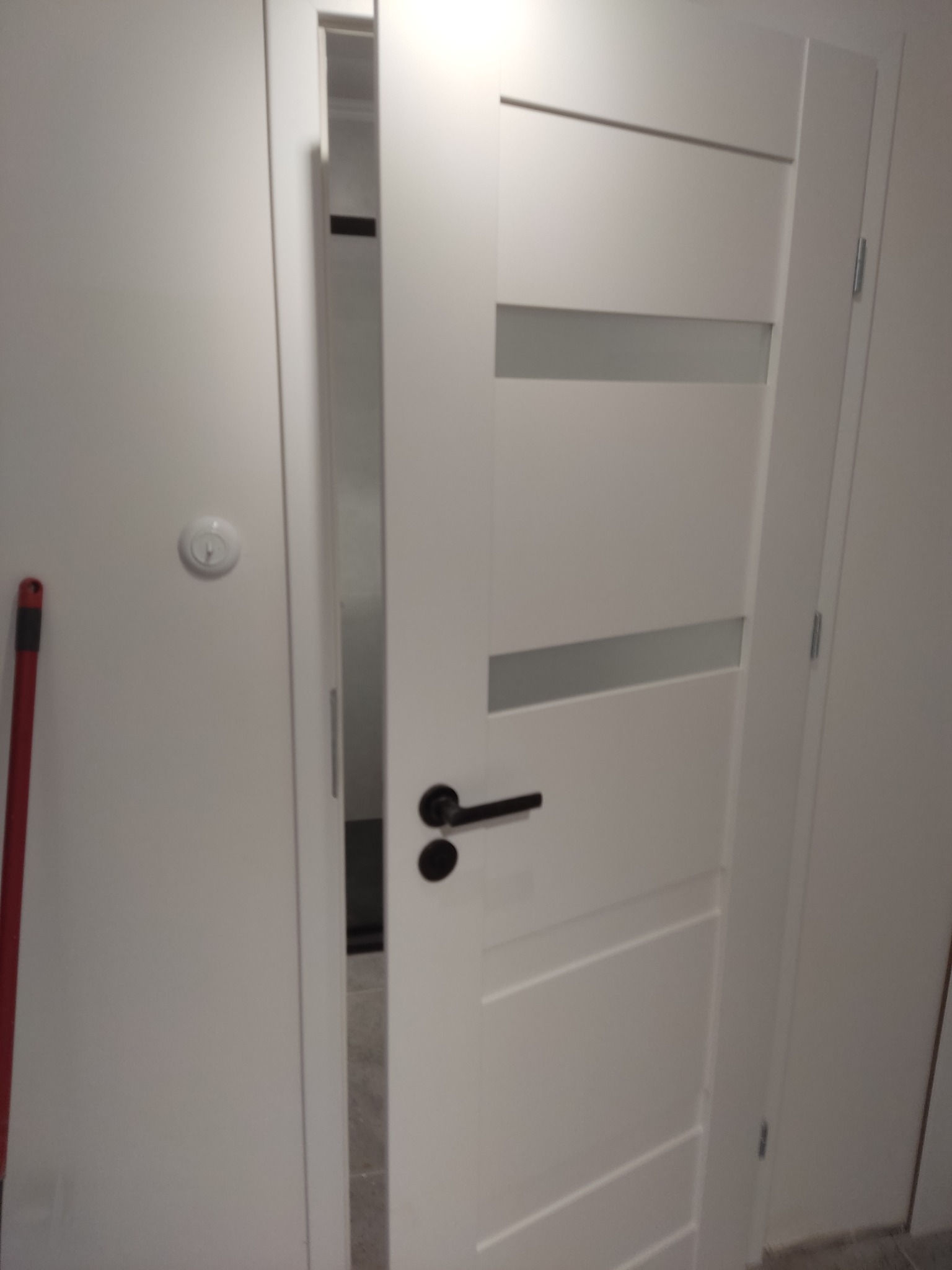 Białe, częściowo otwarte drzwi z przeszkleniami i czarną klamką, widoczne na tle białej ściany z włącznikiem. Czerwony mop oparty o ścianę po lewej stronie.
