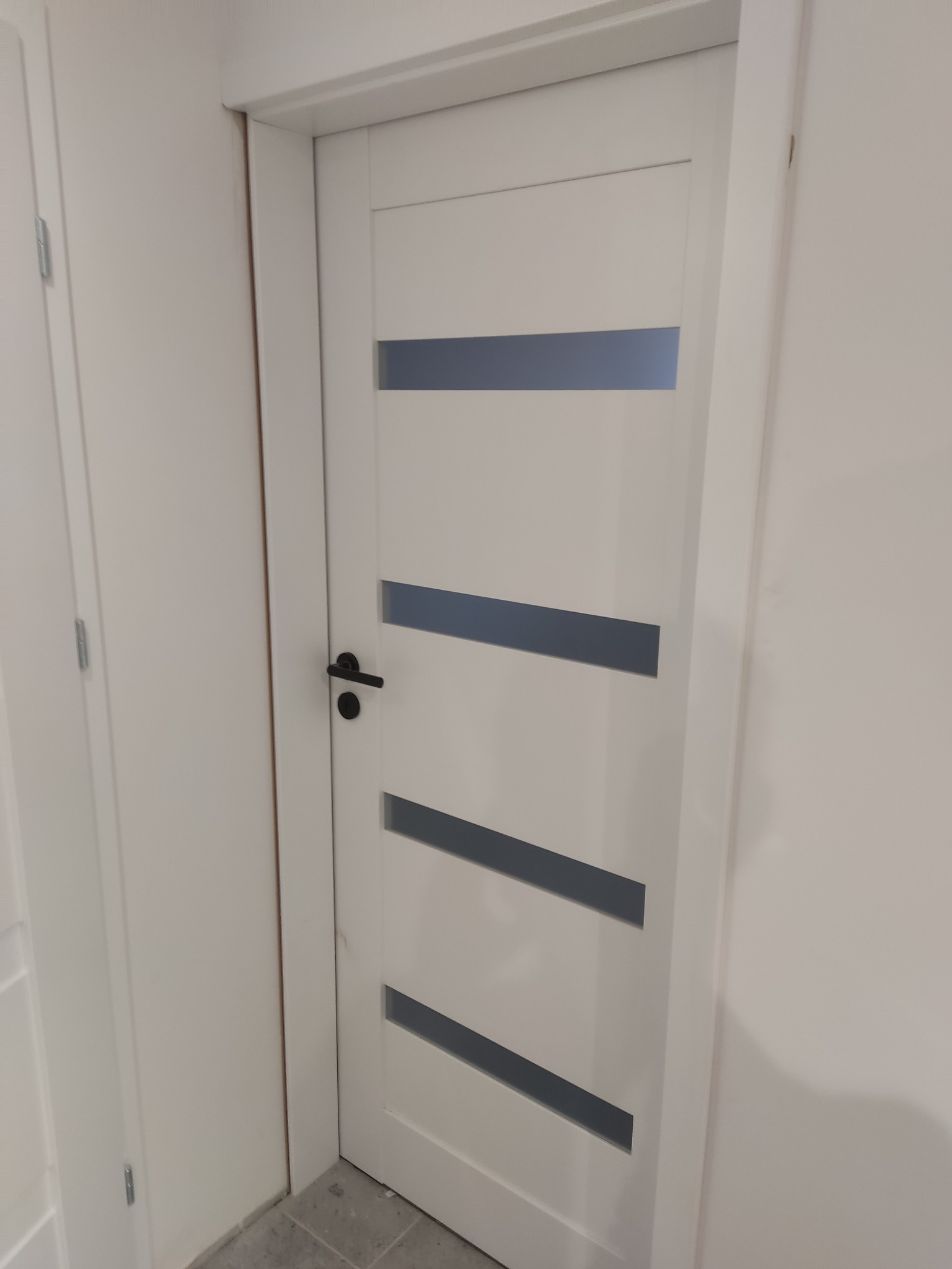 Białe, nowoczesne drzwi wewnętrzne z czterema poziomymi, szarymi przeszkleniami i czarną klamką, zamontowane w białej ścianie. Widoczny fragment podłogi.