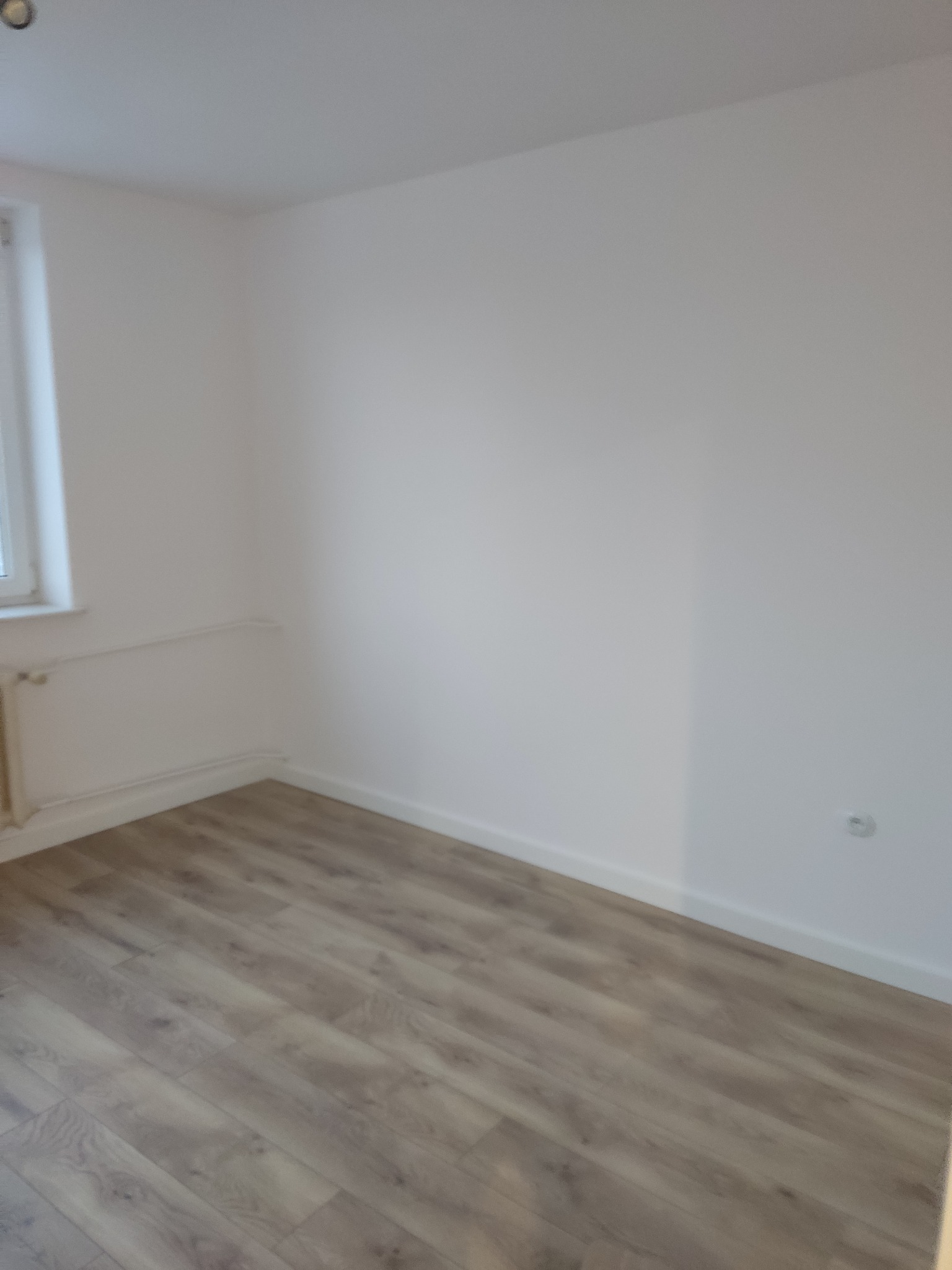 Jasne panele podłogowe w pokoju po remoncie, świeżo pomalowane białe ściany i grzejnik pod oknem. Widoczny kontakt na ścianie.