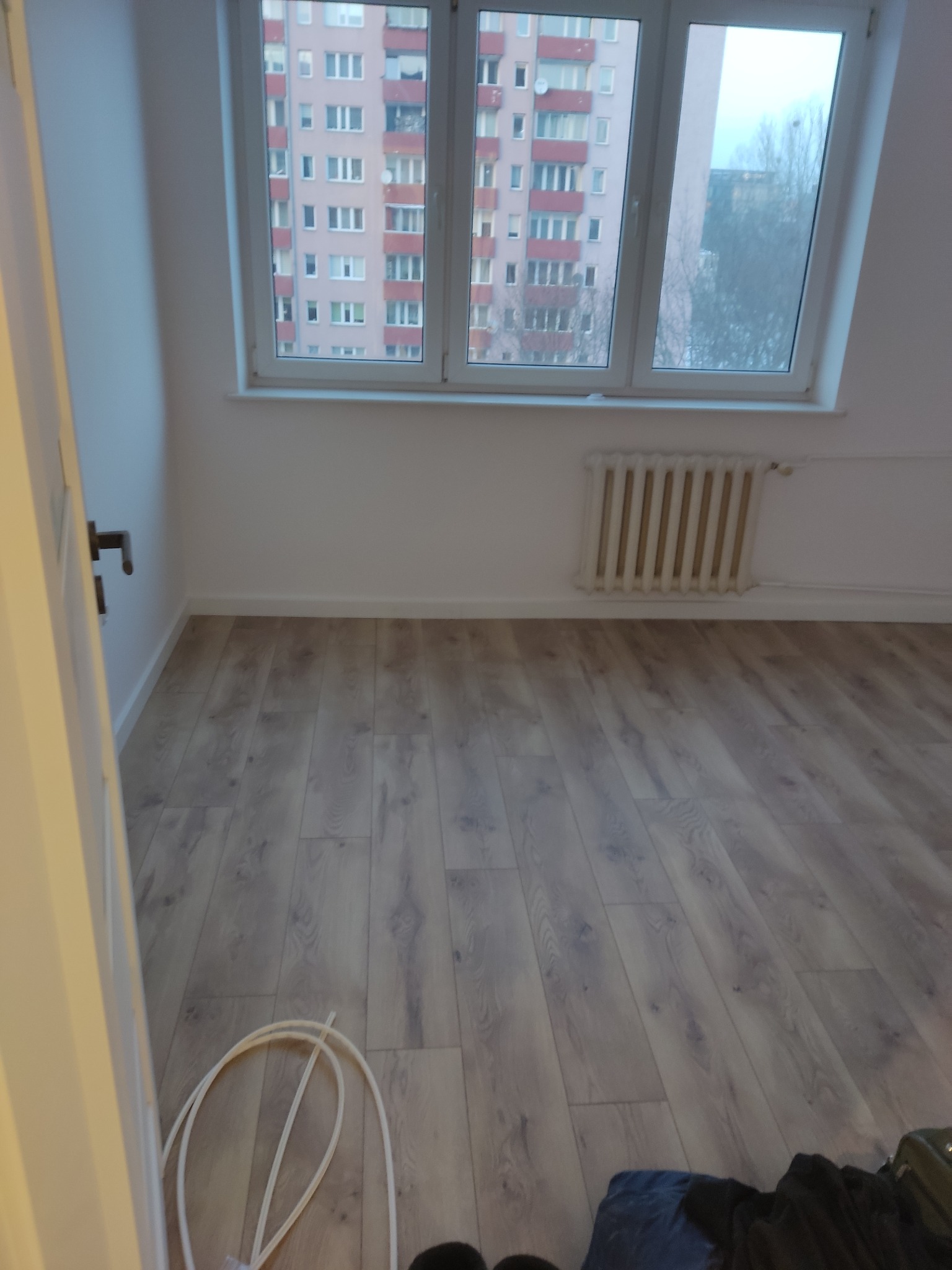 Jasne panele podłogowe w pokoju z grzejnikiem i oknem na blok mieszkalny. Widoczne fragmenty kabli i ubrań. Ujęcie z dołu, z bliska, z otwartych drzwi.