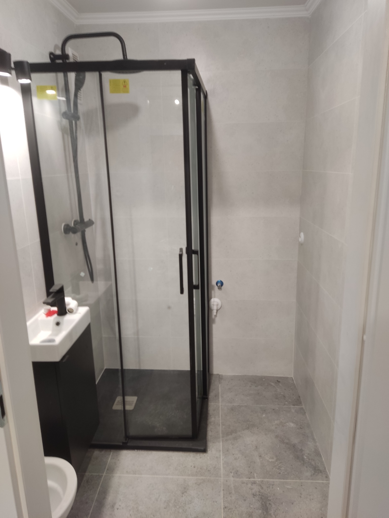 Wykończona łazienka z kabiną prysznicową w czarnej ramie, umywalką i szarą płytką na ścianach i podłodze. Minimalistyczny styl, widoczne elementy instalacji.