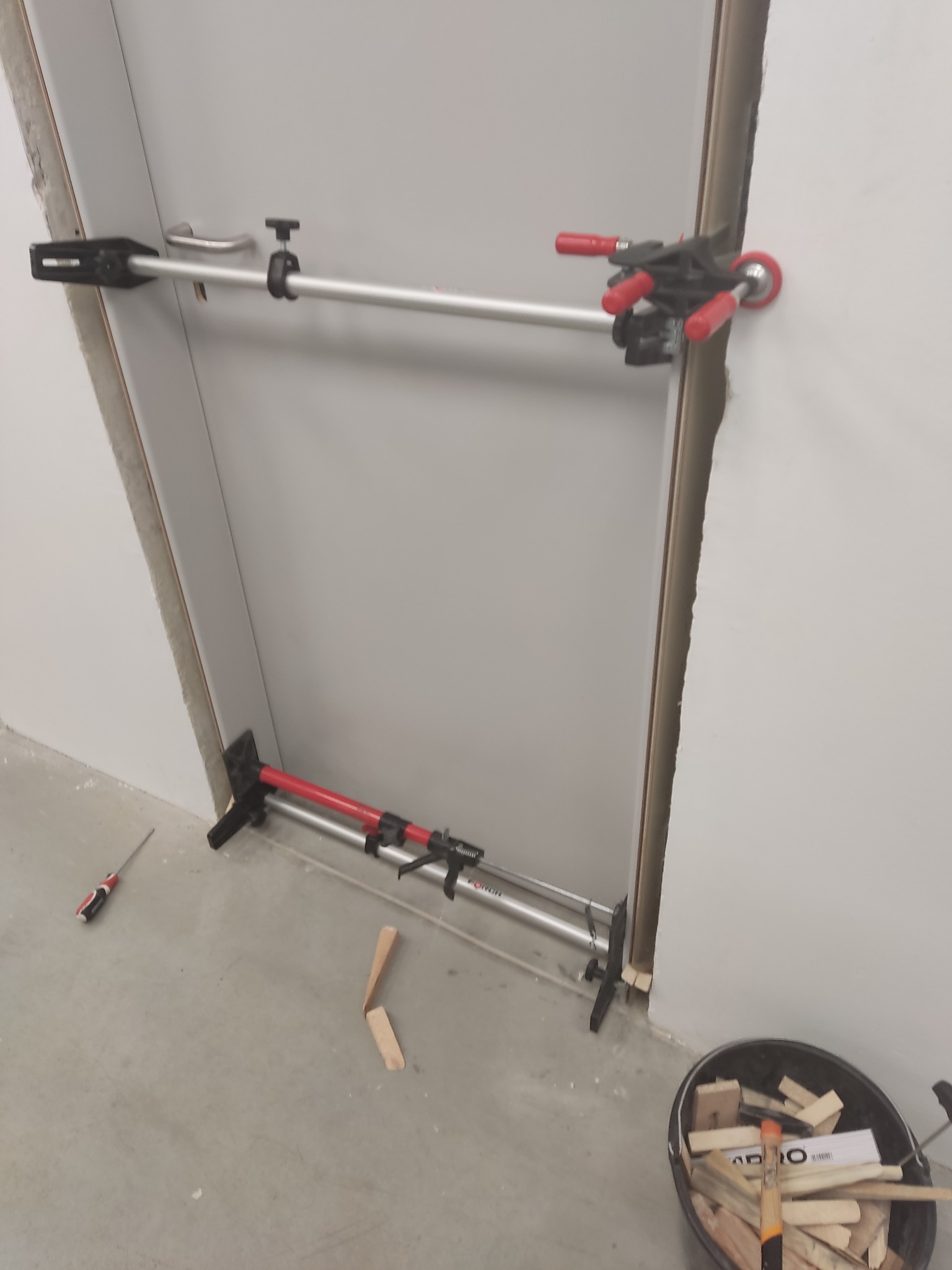 Montaż ościeżnicy drzwiowej za pomocą rozpórki teleskopowej. Widoczne narzędzia i wiadro z drewnianymi elementami. Szara ściana i podłoga w tle.