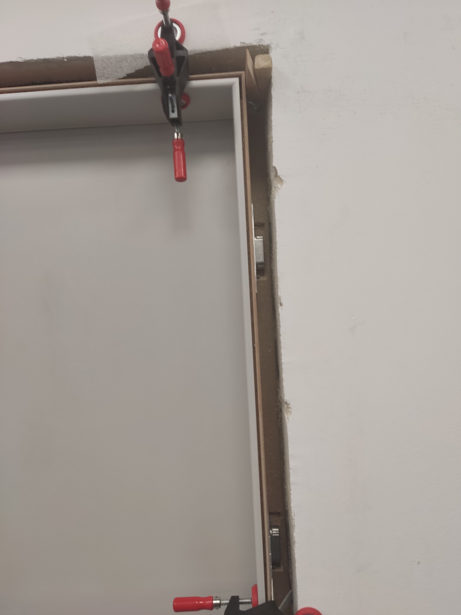 Montaż ościeżnicy z użyciem ścisków stolarskich. Widoczny fragment ściany z uszkodzeniami. Detal z bliska, oświetlenie naturalne, brak dekoracji.