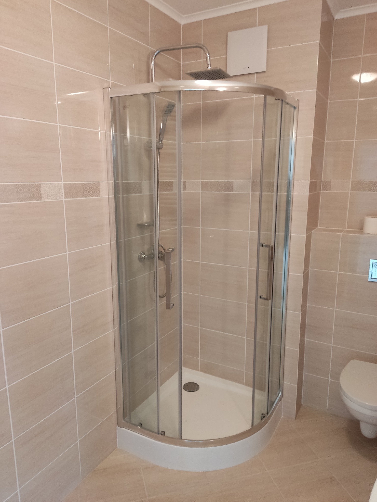 Nowoczesna kabina prysznicowa narożna z chromowanym wykończeniem, beżowe płytki z dekoracyjnym pasem, widoczny fragment toalety.