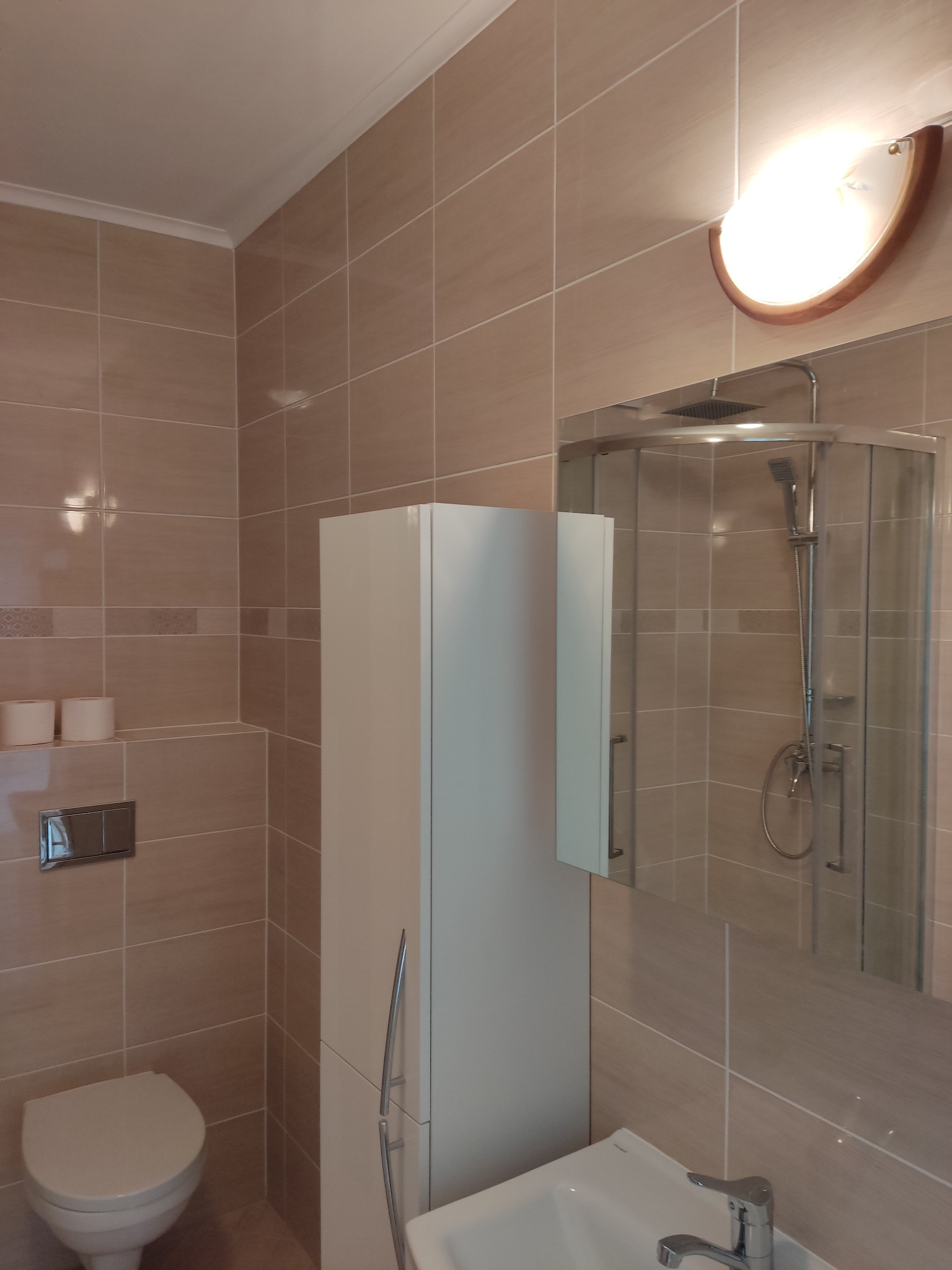 Jasna łazienka z beżowymi płytkami, białą szafką i umywalką. W lustrze odbija się kabina prysznicowa. Widoczne detale: oświetlenie, toaleta.