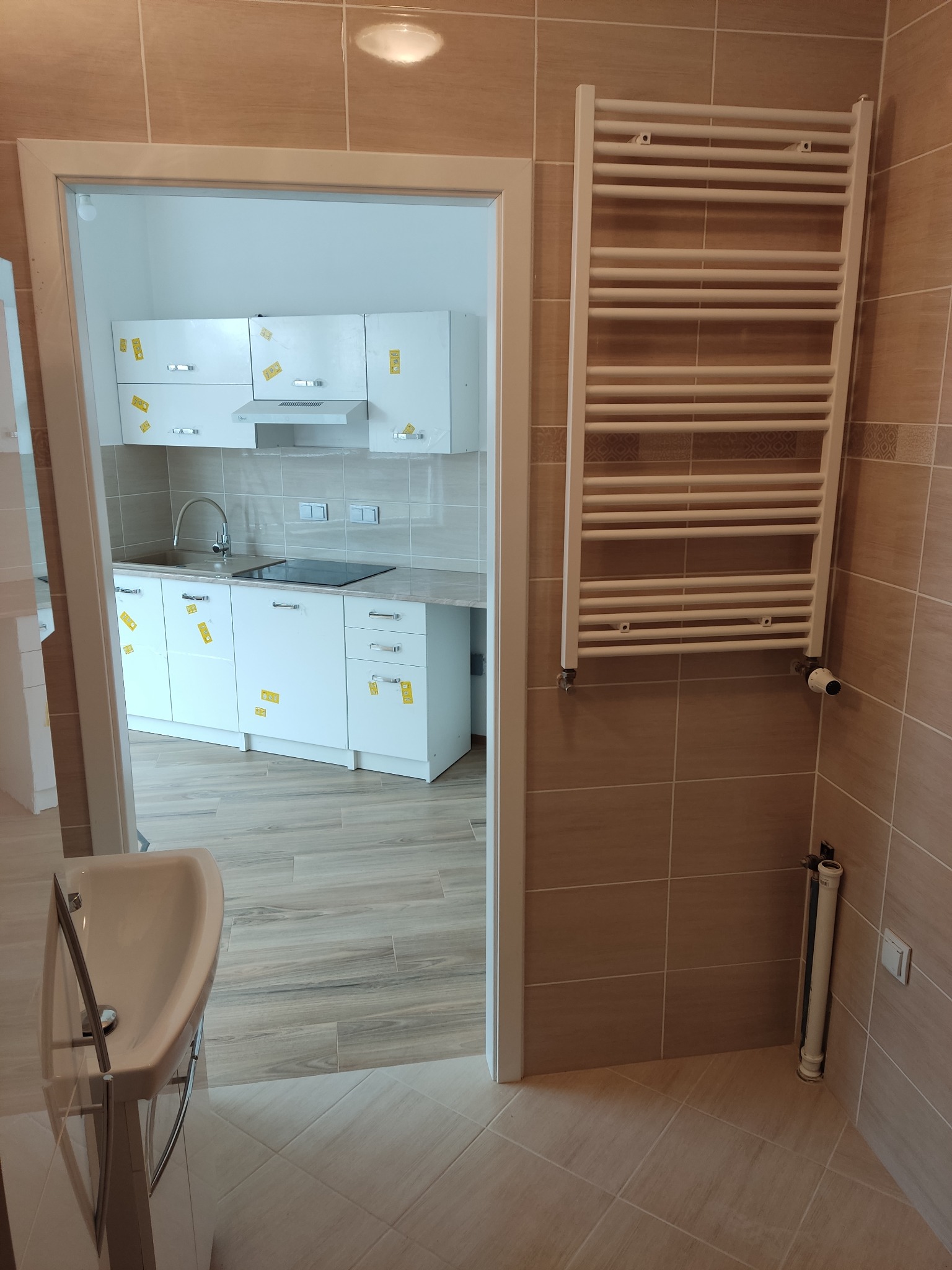 Beżowa łazienka z grzejnikiem drabinkowym i umywalką, widok na białą kuchnię z naklejkami zabezpieczającymi. Całość w jasnych, ciepłych barwach.