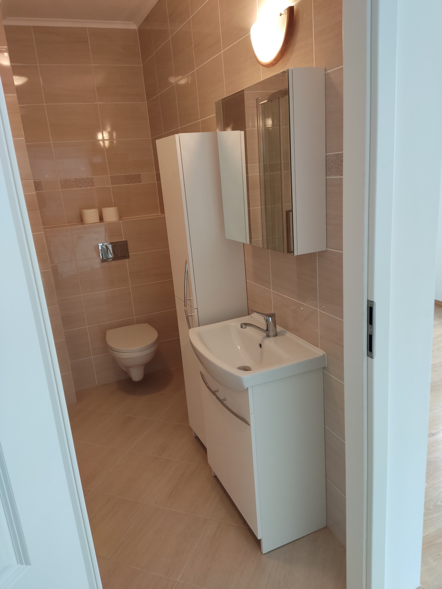 Jasna łazienka z beżowymi płytkami, białą armaturą i szafkami. Widok zza uchylonych drzwi, podkreślony przez ciepłe oświetlenie.