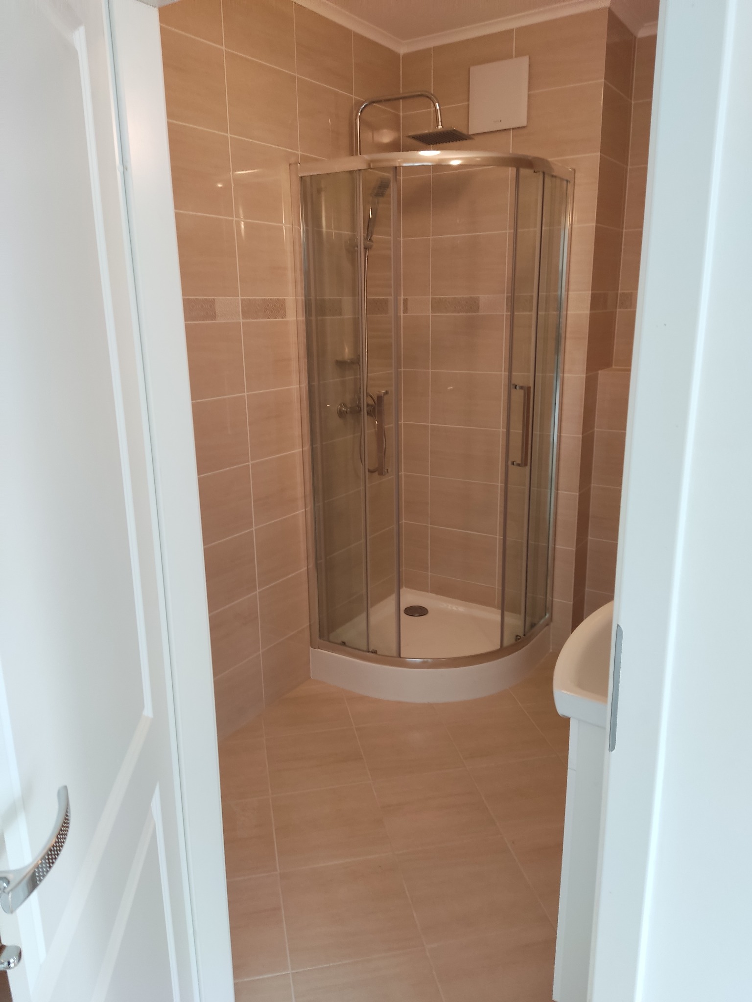 Jasna łazienka z kabiną prysznicową w rogu, wyłożona beżowymi płytkami. Widok przez uchylone białe drzwi. Minimalistyczny design, czysta przestrzeń.